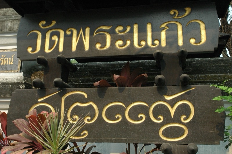 ป้ายไม้สีเข้มสองแผ่นหน้าวัดพระแก้ว เชียงราย ตัวอักษรสีทอง แผ่นบนเป็นอักษรไทย แผ่นล่างเป็นอักษรธรรมล้านนา อ่านว่าวัดพระแก้วเหมือนกัน