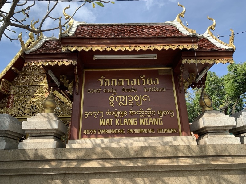 ป้ายหน้าวัดกลางเวียง เชียงราย พื้นสีน้ำตาลแดง ตัวอักษรสีทอง มีชื่อวัดเป็นอักษรไทย อักษรธรรมล้านนา และภาษาอังกฤษ ใต้หลังคาทรงล้านนา