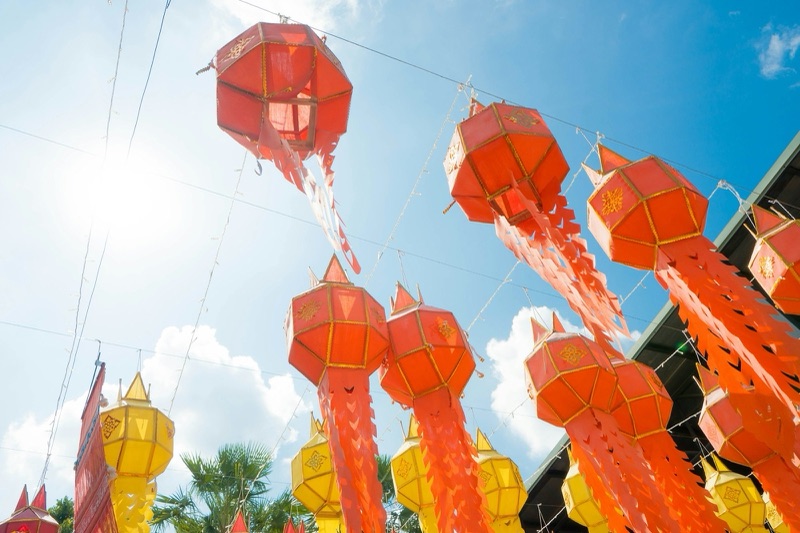โคมกระดาษทรงล้านนาสีแดงและสีเหลืองแขวนเรียงกันใต้ท้องฟ้าสีครามและเมฆขาว มองจากมุมต่ำขึ้นไป