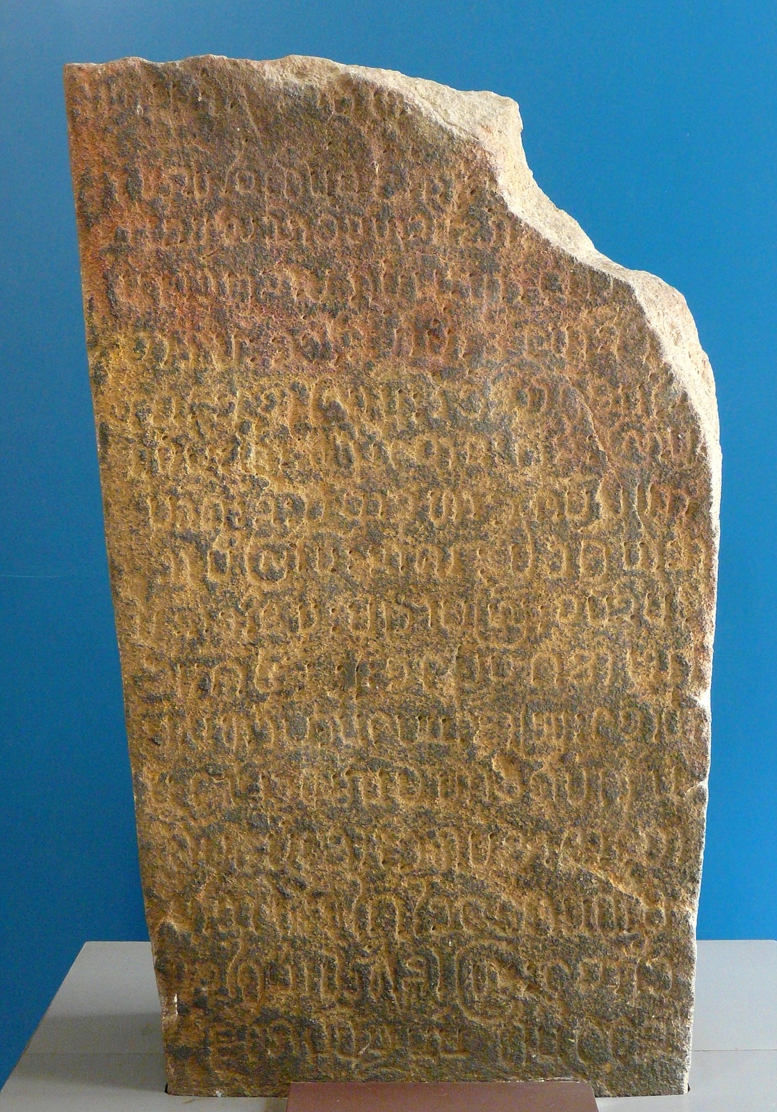 The whole Wat Lam Chang inscription stone, its top broken off at a slant, standing in front of a blue wall in the museum