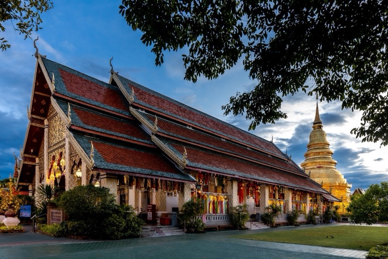 วิหารหลวงวัดพระธาตุหริภุญชัย ลำพูน หลังคาซ้อนชั้นสีแดง มีพระบรมธาตุสีทองอยู่ด้านหลัง ยามพลบค่ำ