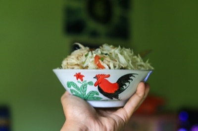 A hand holding a white bowl painted with a red and black rooster and a green banana plant