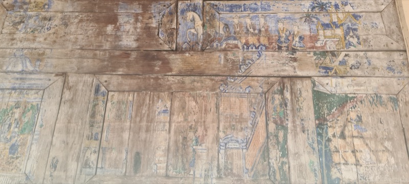 A wooden panel in the main vihara of Wat Phra That Lampang Luang with faded paintings of figures and buildings