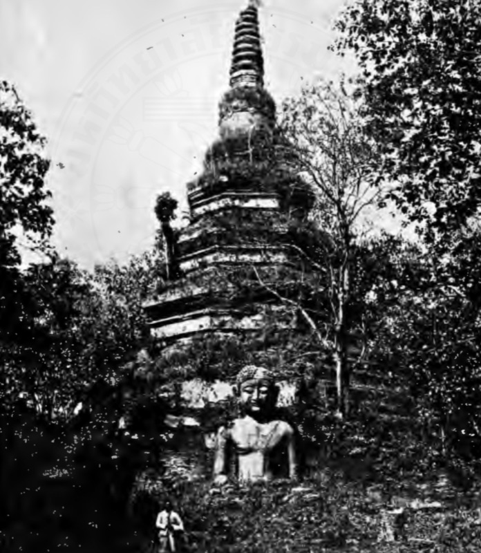 An old black-and-white photograph of the tall brick chedi of Wat Phra Chedi Luang, Chiang Saen, overgrown with trees, with a Buddha image in front