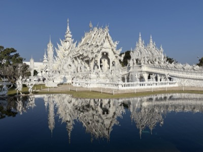 The white ordination hall of Wat Rong Khun with intricate decoration, reflected in a pond under a deep blue sky