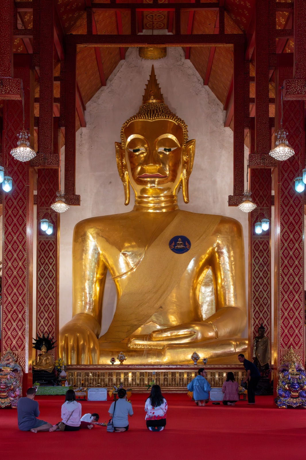 A huge golden seated Buddha subduing Mara in a hall with red and gold pillars; people sit below with their backs to the camera, tiny next to the image