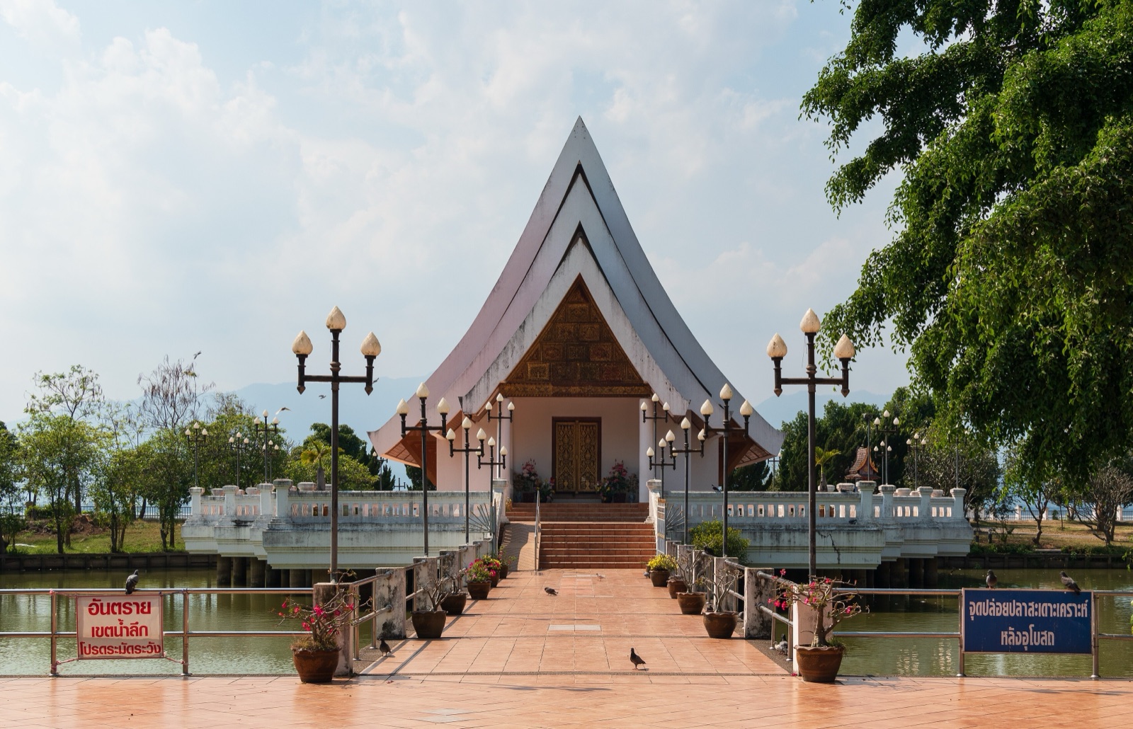 A white ordination hall with tall tiered roofs standing in a pond, a walkway lined with ornamental lamp posts leading to its front stairs