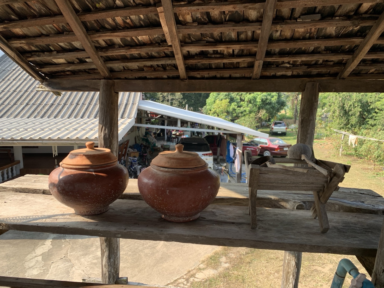 Two lidded red clay water jars on a wooden shelf under the eaves of a wooden house, a wooden ladle beside them, a yard and a car behind