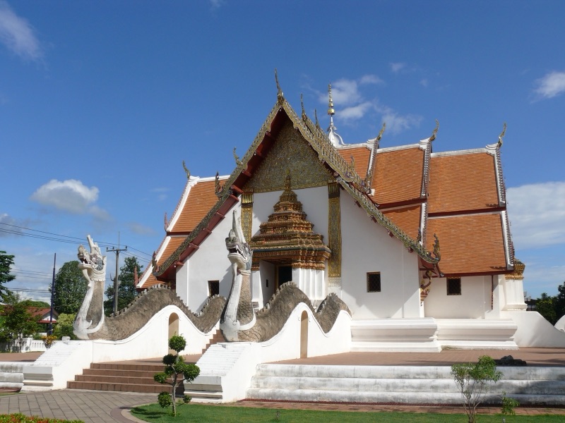 วิหารวัดภูมินทร์ เมืองน่าน ผนังสีขาว หลังคากระเบื้องสีส้มซ้อนชั้น หน้าบันสีทอง มีพญานาคคู่ทอดตัวเป็นราวบันไดทั้งสองข้าง ใต้ท้องฟ้าสีฟ้า