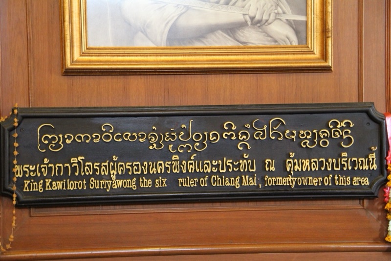 ป้ายไม้สีดำตัวอักษรสีทอง บรรทัดบนเป็นอักษรธรรมล้านนา บรรทัดล่างเป็นอักษรไทยและอังกฤษ ติดอยู่ใต้กรอบรูปสีทอง