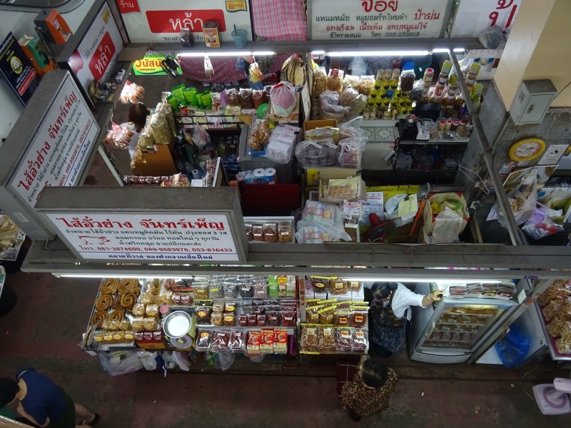 มองจากชั้นบนลงไปยังร้านขายของฝากในตลาดวโรรส เชียงใหม่ ป้ายร้านภาษาไทยสีแดงขาวเขียนชื่อร้านและสินค้า มีของกินของฝากวางเรียงเต็มร้าน