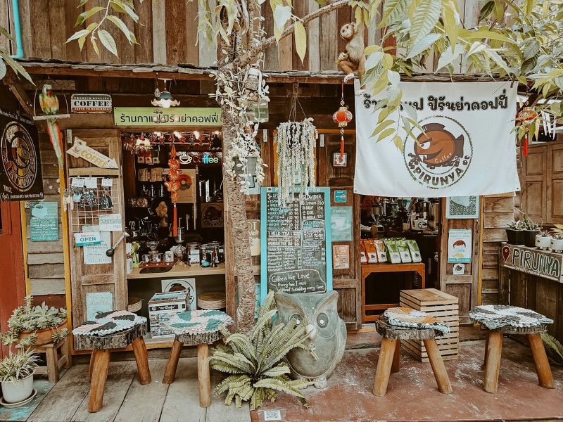 หน้าร้านกาแฟไม้เล็ก ๆ ในหมู่บ้านบนดอยของเชียงใหม่ มีม้านั่งไม้หน้าร้าน กระดานเมนู และต้นไม้ประดับ