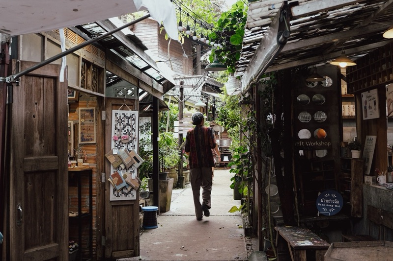 คนเดินหันหลังไปตามตรอกแคบ ๆ ที่มีร้านไม้เก่าเรียงสองข้าง ต้นไม้และไฟห้อยอยู่เหนือทางเดิน