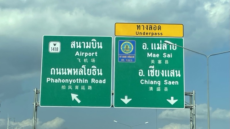 Green road signs in Chiang Rai reading Airport, Phahonyothin Road, Mae Sai District and Chiang Saen District, with a yellow Underpass sign above