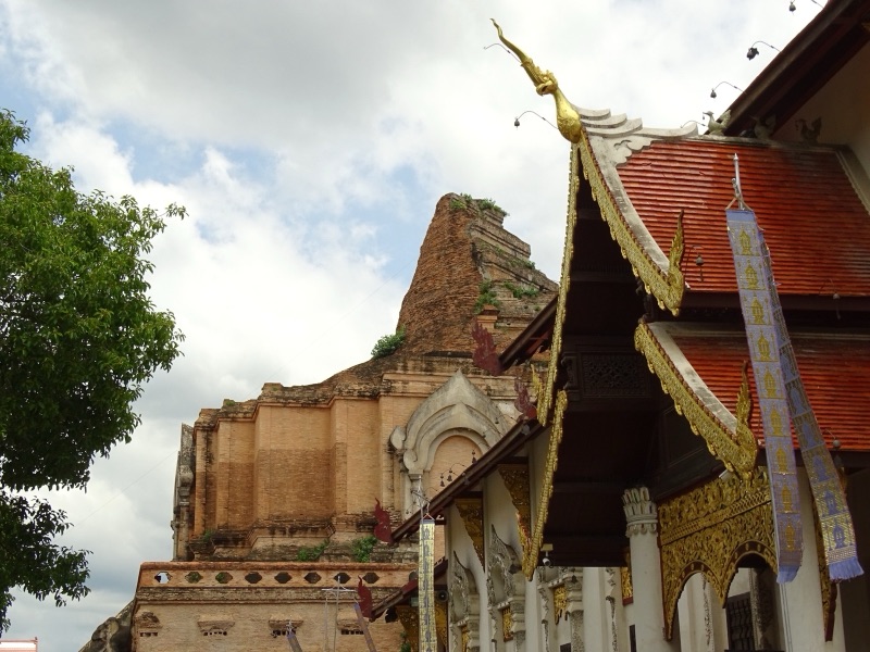 ซากพระเจดีย์หลวงเชียงใหม่ องค์เจดีย์อิฐขนาดใหญ่ที่ส่วนยอดพังหายไป ด้านหน้ามีหน้าบันวิหารสีแดงทองใต้ท้องฟ้ามีเมฆ