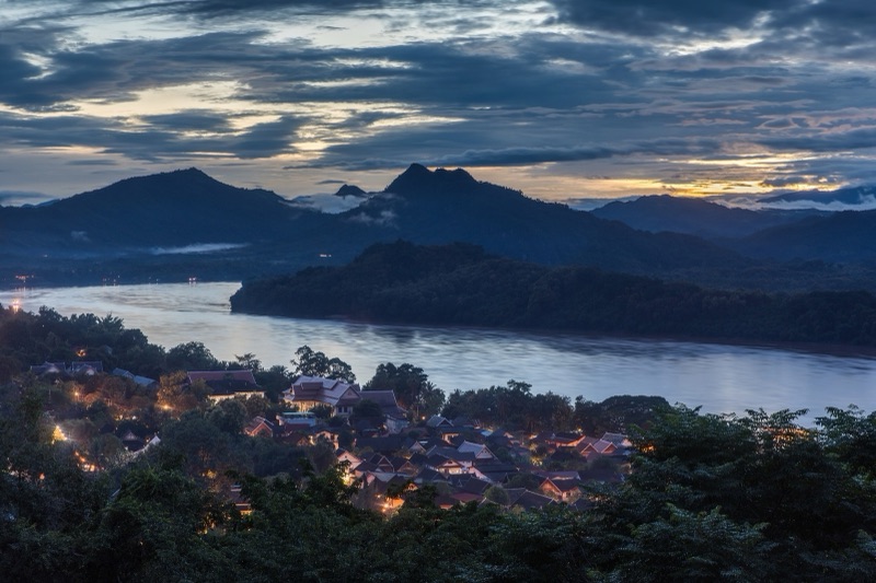 The wide Mekong flowing past Luang Prabang at dusk, with houses on the bank and mountains behind