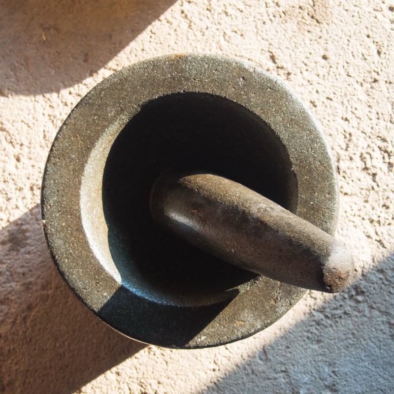 A dark grey stone mortar with its stone pestle resting inside, seen from above on a sunlit concrete floor