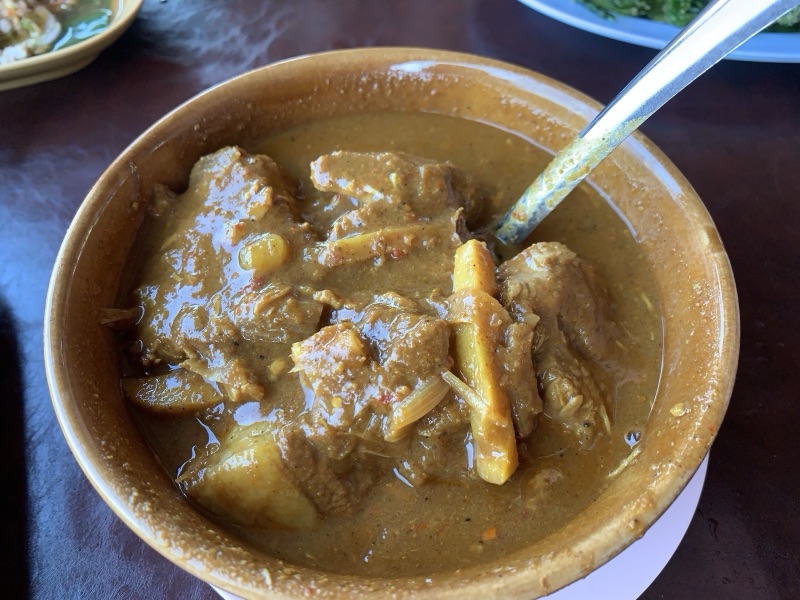 Hang le curry in an earthenware bowl with large pieces of pork and sticks of ginger, a spoon resting in the bowl, on a dark table