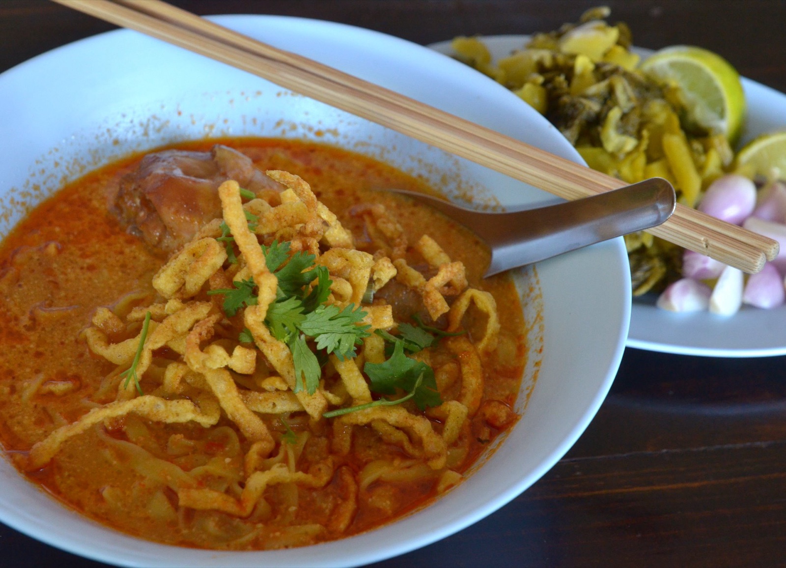Chicken khao soi in a white bowl with a drumstick and crispy noodles, a plate of pickled mustard greens, shallots and lime behind it