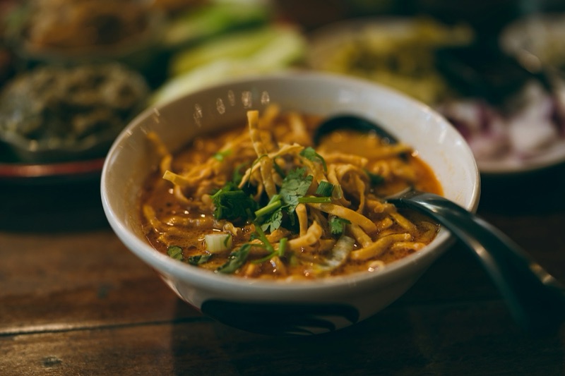 Orange khao soi curry in a white bowl topped with crispy noodles and coriander, a spoon in the bowl, on a wooden table with side dishes