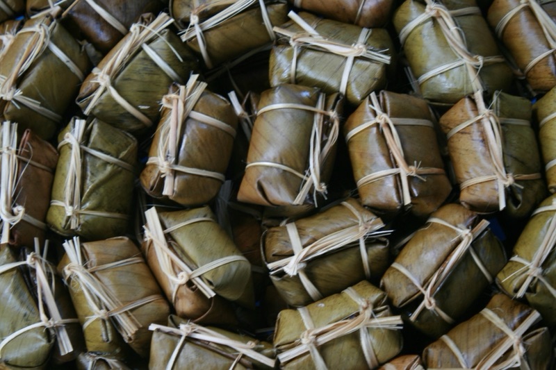 A pile of khao tom mat, sticky rice parcels wrapped in banana leaf and tied with string, in green and golden yellow