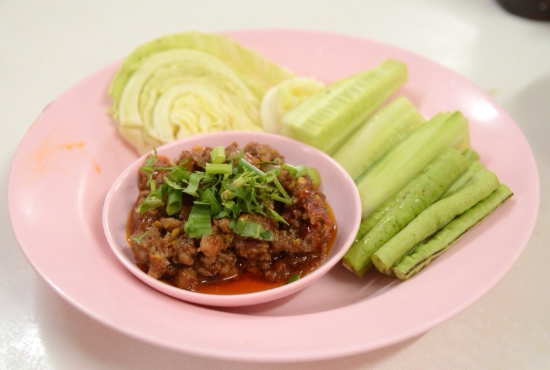 Orange nam phrik ong in a small bowl topped with spring onion, on a pink plate with cabbage and cucumber sticks