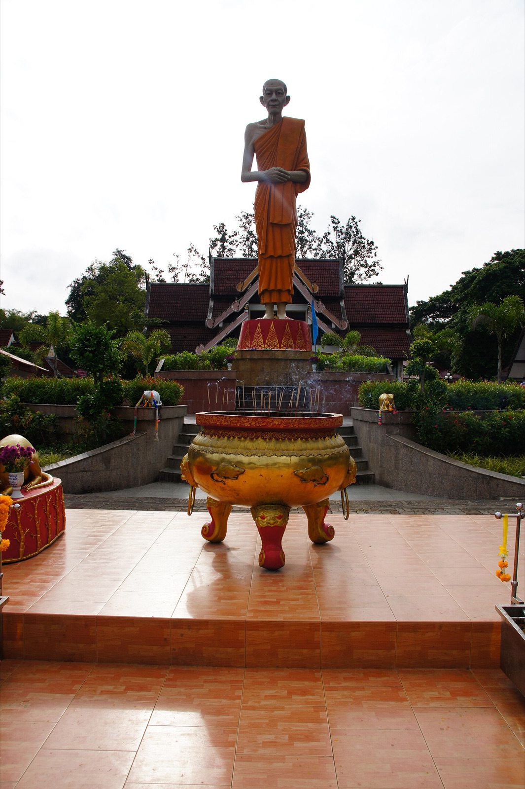 รูปปั้นพระภิกษุครองจีวรสีส้มยืนบนแท่นสูง ด้านหน้ามีกระถางธูปสีทองขนาดใหญ่