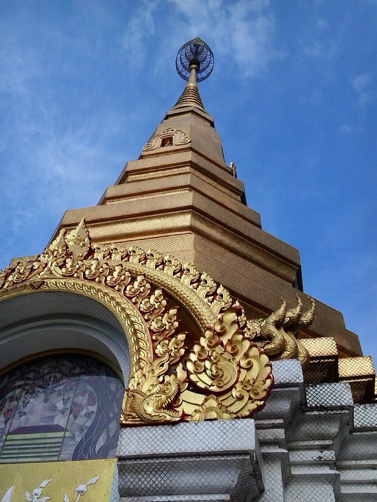 ยอดพระธาตุเจดีย์สีทองทรงเหลี่ยมซ้อนชั้น ซุ้มประตูประดับลายนาคสีทอง ตัดกับท้องฟ้าสีคราม