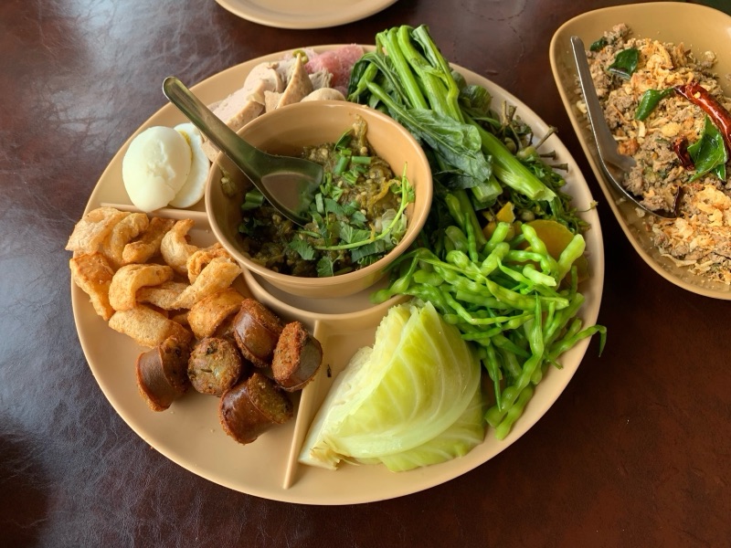อาหารเมืองจัดเป็นสำรับ มีน้ำพริกหนุ่ม แคบหมู ไส้อั่ว ไข่ต้ม และผักสดผักลวกวางรวมในถาดเดียว