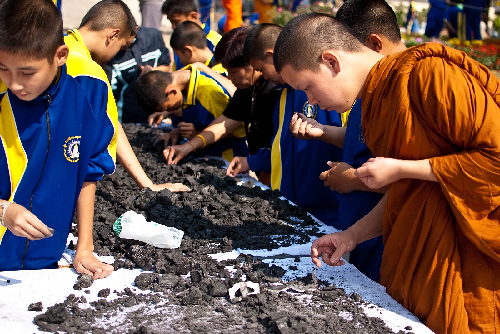 A monk and students bending over ashes spread on white cloth, looking for relics