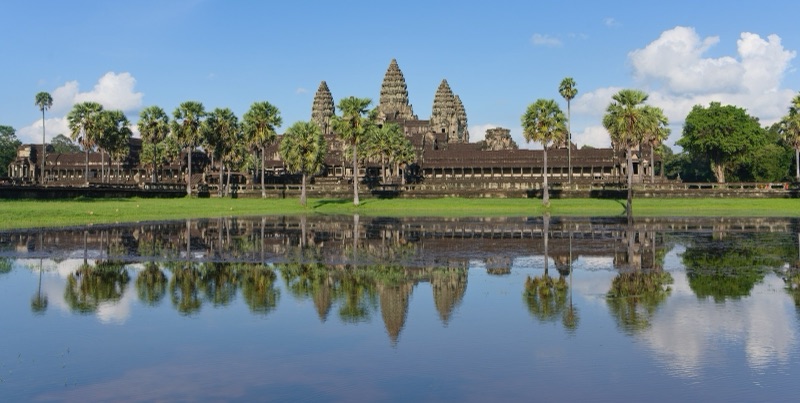 ปราสาทนครวัดห้ายอดสะท้อนในสระน้ำหน้าปราสาท มีต้นตาลเรียงรายใต้ท้องฟ้าสีฟ้าและเมฆขาว