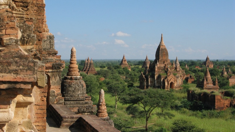 The plain of Bagan in Myanmar, with many ancient brick stupas and temples scattered among green trees under a blue sky