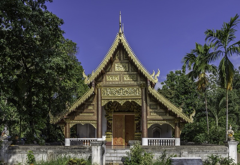 The Lanna-style assembly hall of Wat Chiang Man in Chiang Mai, with tiered roofs and gold decoration, among trees and palms under a deep blue sky