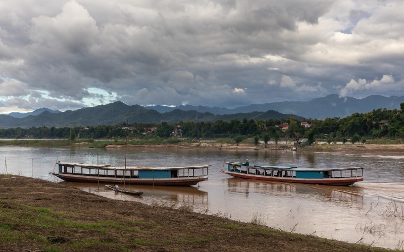 แม่น้ำโขงที่หลวงพระบางยามบ่าย มีเรือไม้ลำยาวจอดริมฝั่ง ทิวเขาสีเขียวเข้มอยู่ไกล และเมฆฝนสีเทาลอยเต็มฟ้า