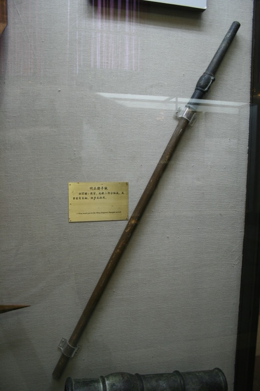 A small bronze gun with a long barrel and a hollow socket at the back for a wooden pole, displayed in a museum case