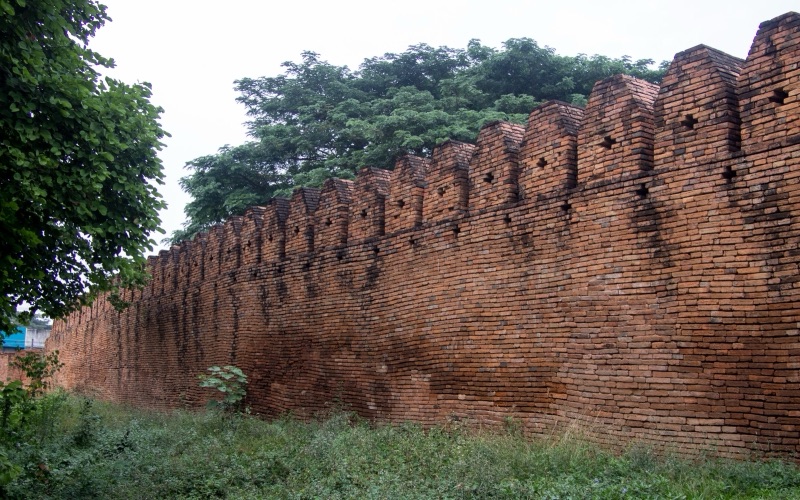 A stretch of old orange-red brick city wall with battlements along the top, a green lawn and large trees in front
