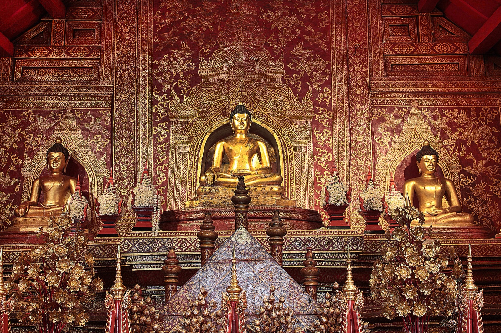 พระพุทธรูปสีทองปางมารวิชัยสามองค์ประดิษฐานหน้าผนังลายทองบนพื้นแดง องค์กลางใหญ่ที่สุด ด้านหน้ามีเครื่องสักการะ