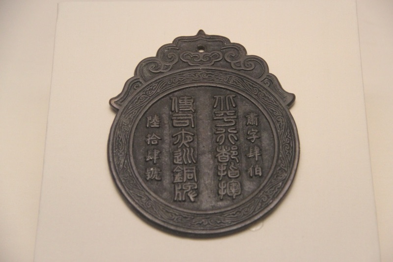 A dark round copper tablet with a cloud-shaped loop on top, raised old Chinese characters in vertical lines, displayed in a museum case