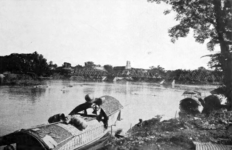 Old black-and-white photograph of the wide Ping River, a boat with a curved roof moored in front and an iron truss bridge across the river in the distance
