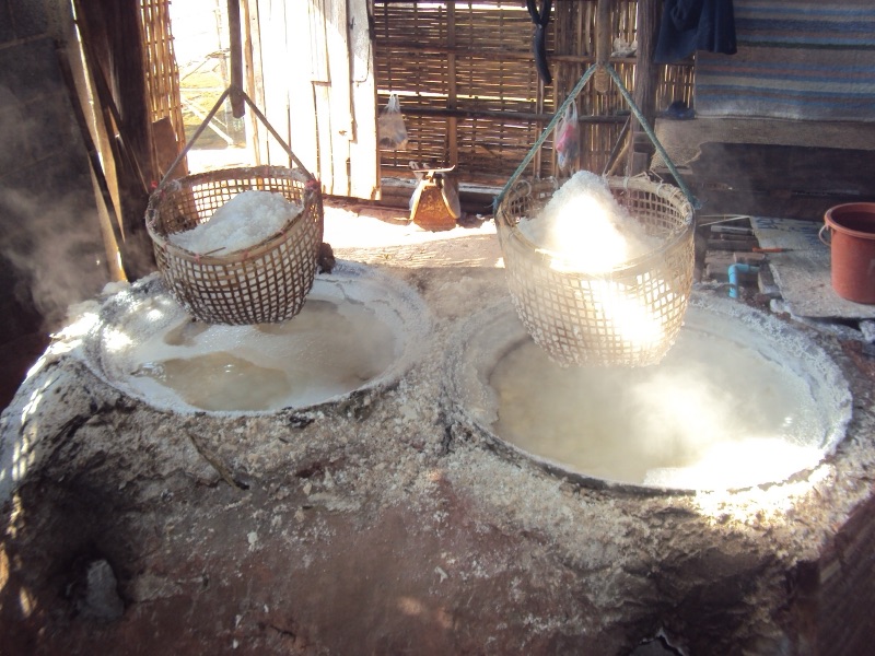 Two large pans of brine boiling on an earthen stove, steam rising, with bamboo baskets of white salt hanging above them inside a bamboo shed