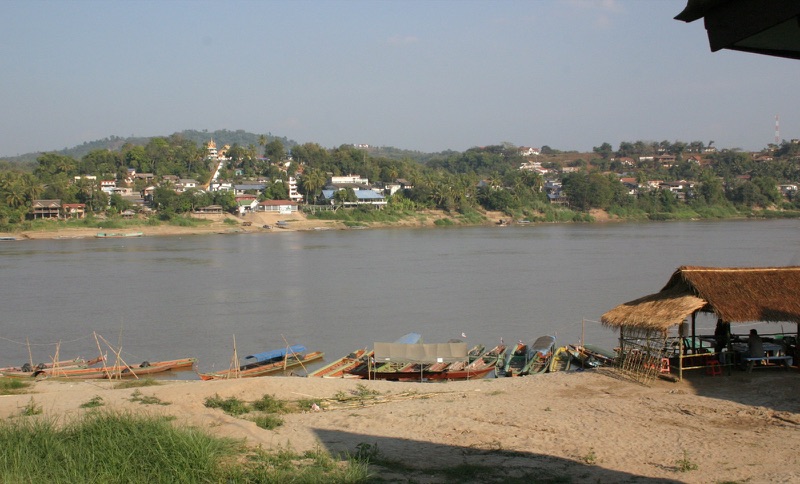 The Mekong at Chiang Khong, with long-tail boats moored along a sandy bank and a thatched shelter; across the river, the Lao town of Huay Xai on a hillside