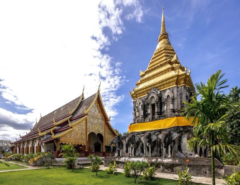 วัดเชียงมั่น เชียงใหม่ วิหารหน้าบันปิดทองกับเจดีย์ที่มีรูปช้างล้อมรอบฐาน ใต้ท้องฟ้าสีคราม