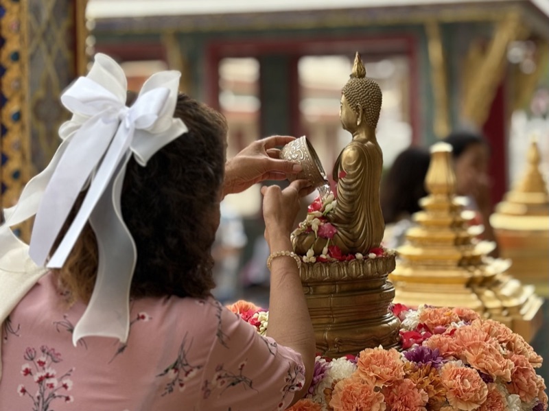 A woman with a white bow pours water over a flower-decked Buddha image at Songkran, seen from behind