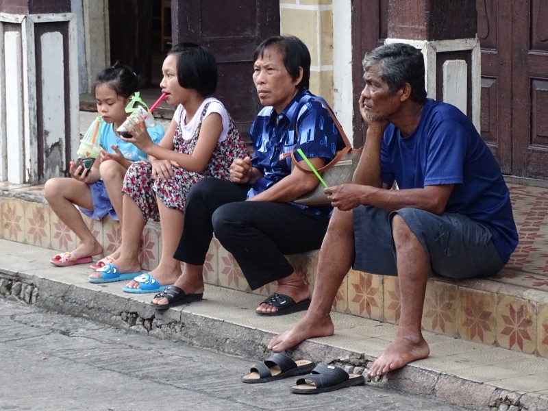 Two adults and two children resting on the front step of a house by the street in Lampang