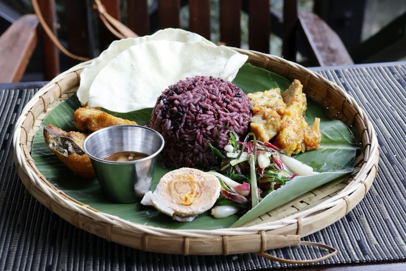 สำรับอาหารบนถาดสานรองใบตอง มีข้าวสีม่วง ไข่ต้ม ของทอด ผักสด และถ้วยน้ำจิ้ม