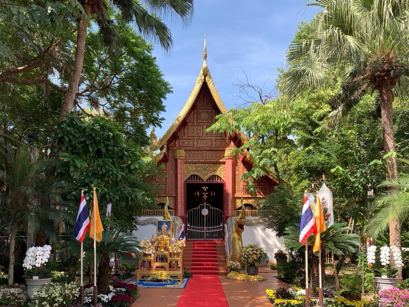 อุโบสถวัดพระแก้ว เชียงราย หน้าบันไม้แกะสลักสีแดงทอง ทางเดินพรมแดงขนาบด้วยธงชาติ ธงเหลือง และต้นไม้ร่มรื่น