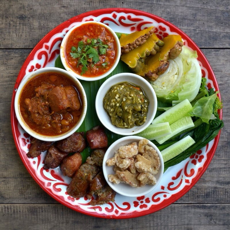 ถาดอาหารเหนือ มีน้ำพริกอ่อง น้ำพริกหนุ่ม แกงฮังเล ไส้อั่ว แคบหมู และผักสดวางบนใบตอง