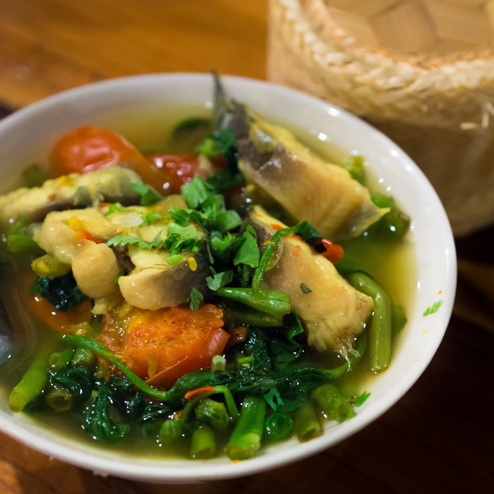 Water spinach and fish curry in a white bowl, with tomato, coriander and pieces of fish, and a sticky-rice basket behind