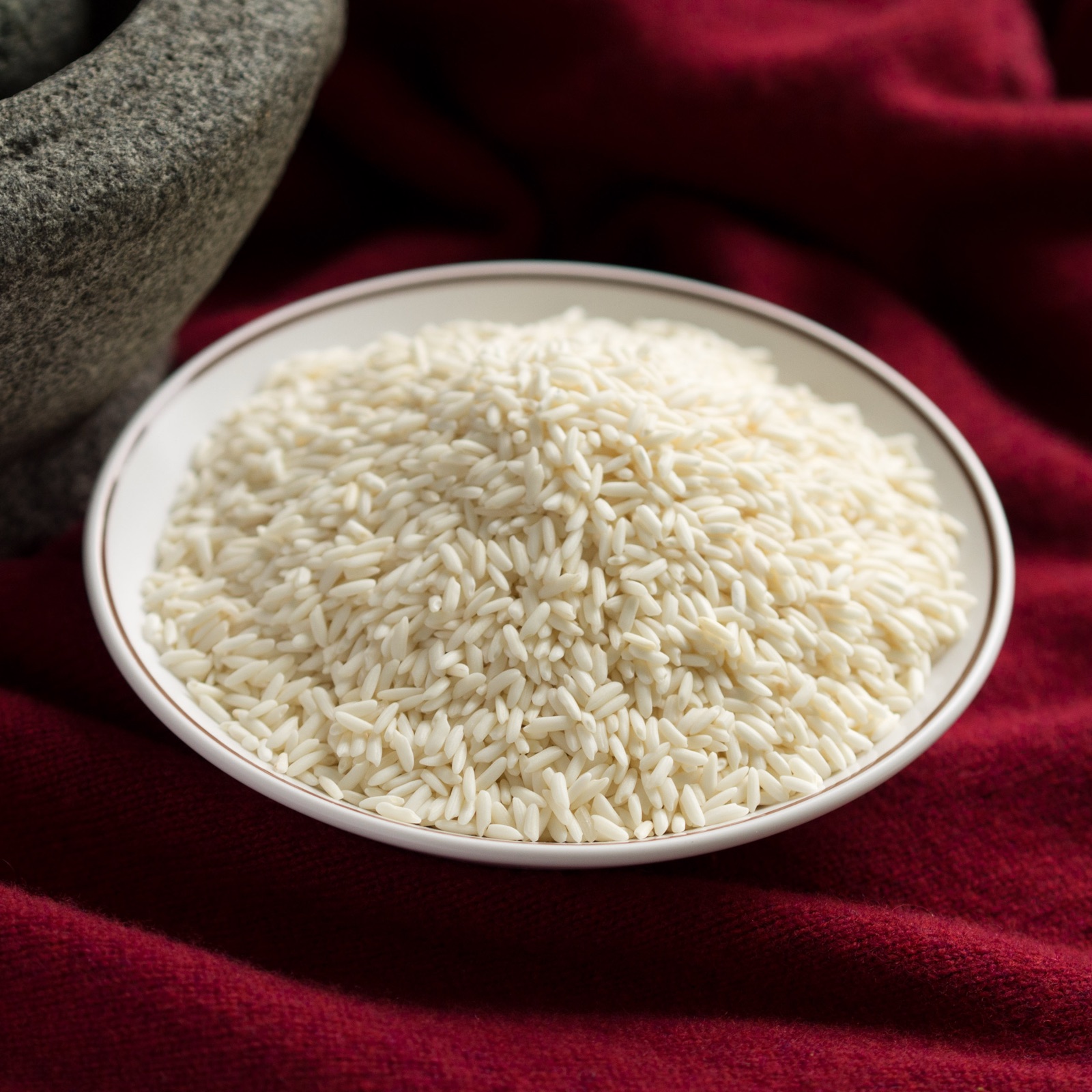 A pile of opaque white glutinous rice grains on a white plate, with a stone mortar beside it on red cloth