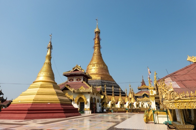 Two golden stupas and a tiered-roof building at Kyaikthanlan Pagoda in Mawlamyine, Myanmar, under a blue sky
